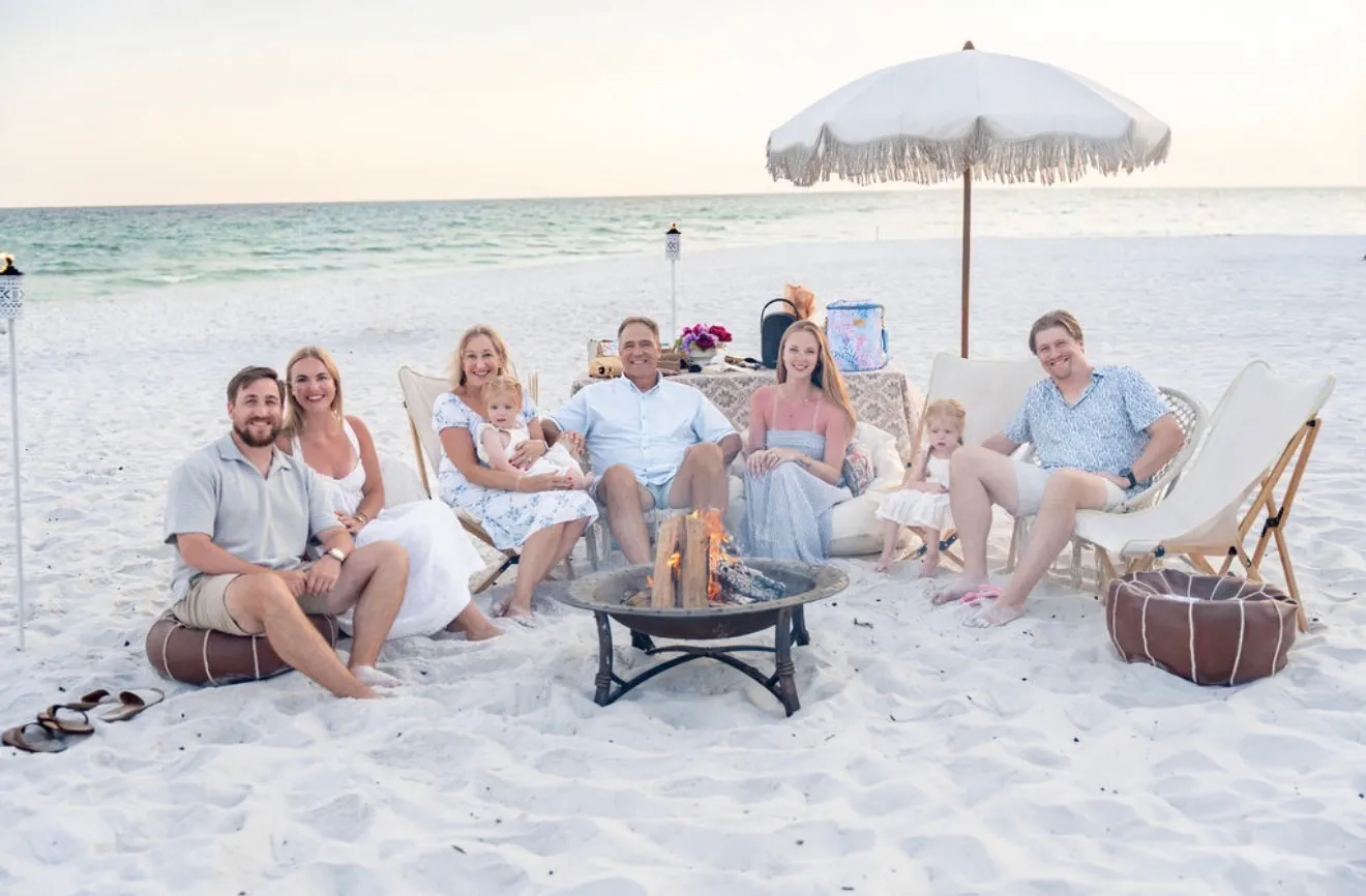 Three generations of a family gathered around a beach bonfire at sunset, smiling together for a photo