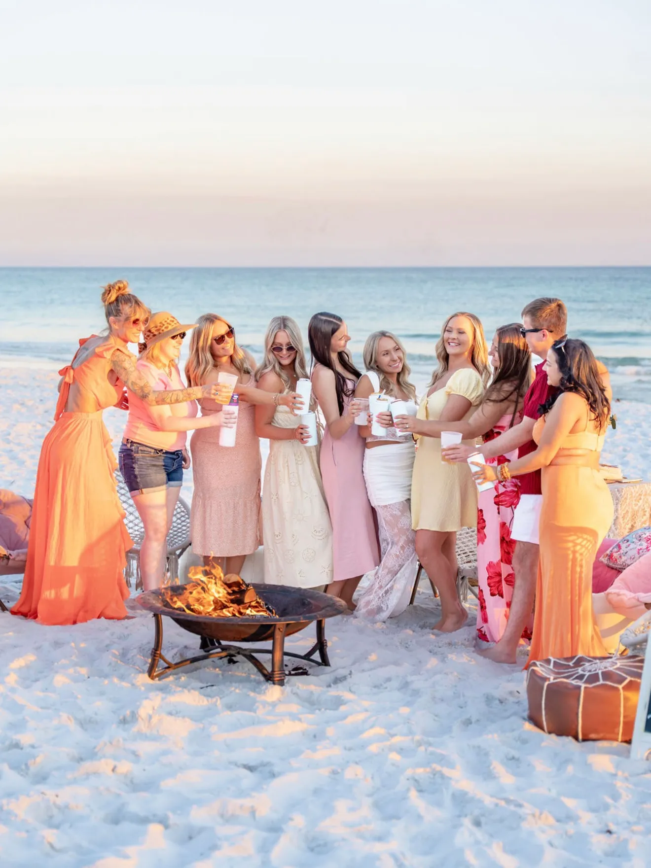 A group of friends in pastel dresses toasting with drinks around the beach bonfire at sunset