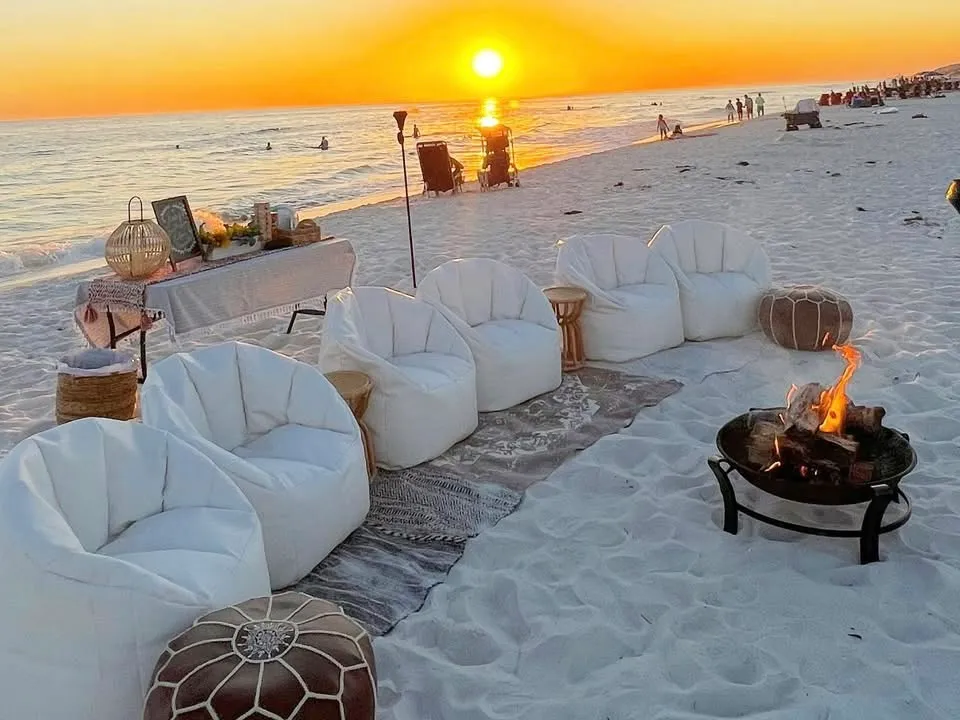 Empty lounge chairs and a glowing fire pit set up on the beach as the sun sets over the water