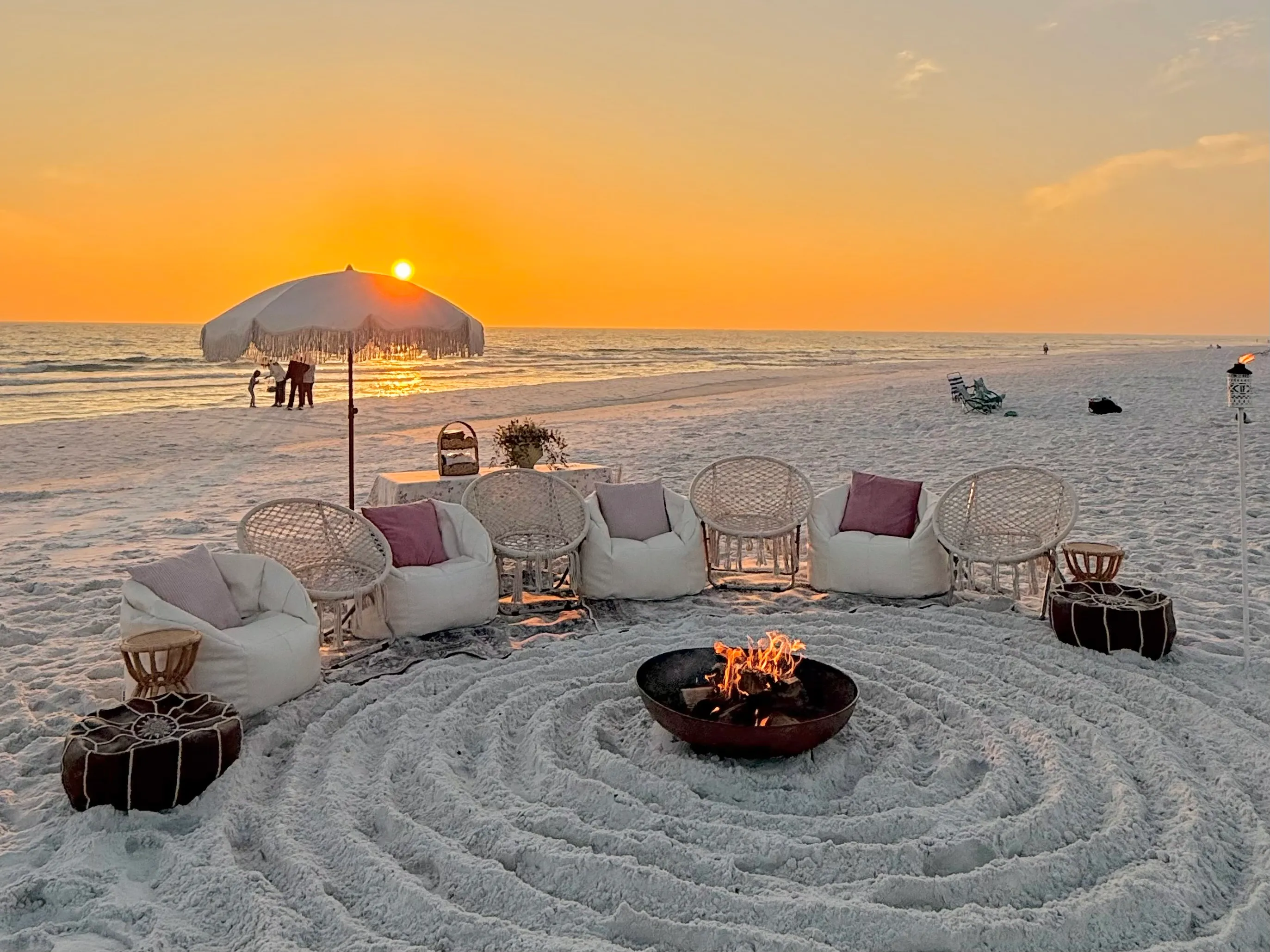 A private beach bonfire lounge setup on 30A with white bean bag chairs and a fringed umbrella as the sun sets over the Gulf
