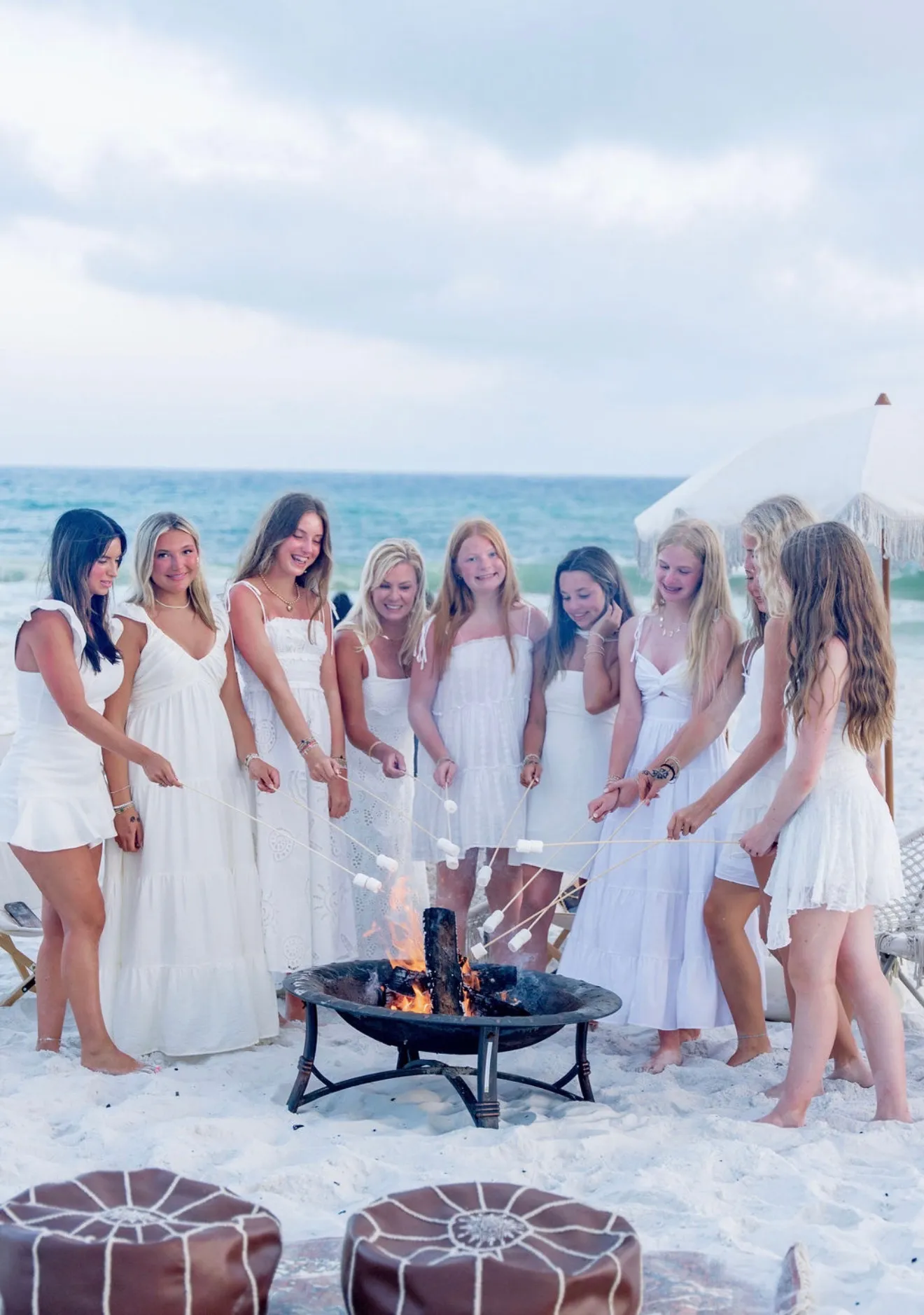 A group of young women in white dresses roasting marshmallows together over a beach bonfire