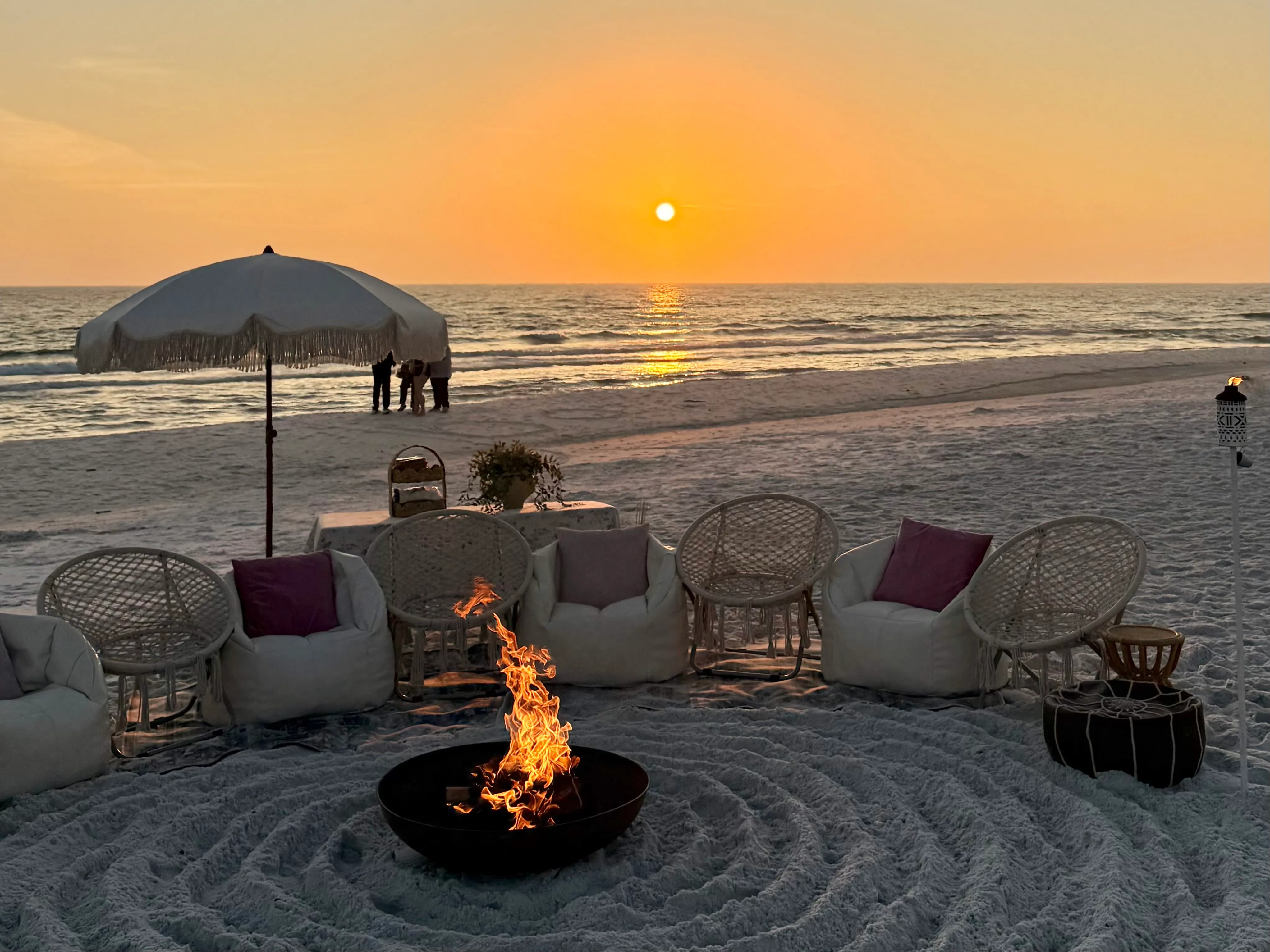 A beach bonfire setup with lounge chairs and a blazing fire pit glowing against the sunset