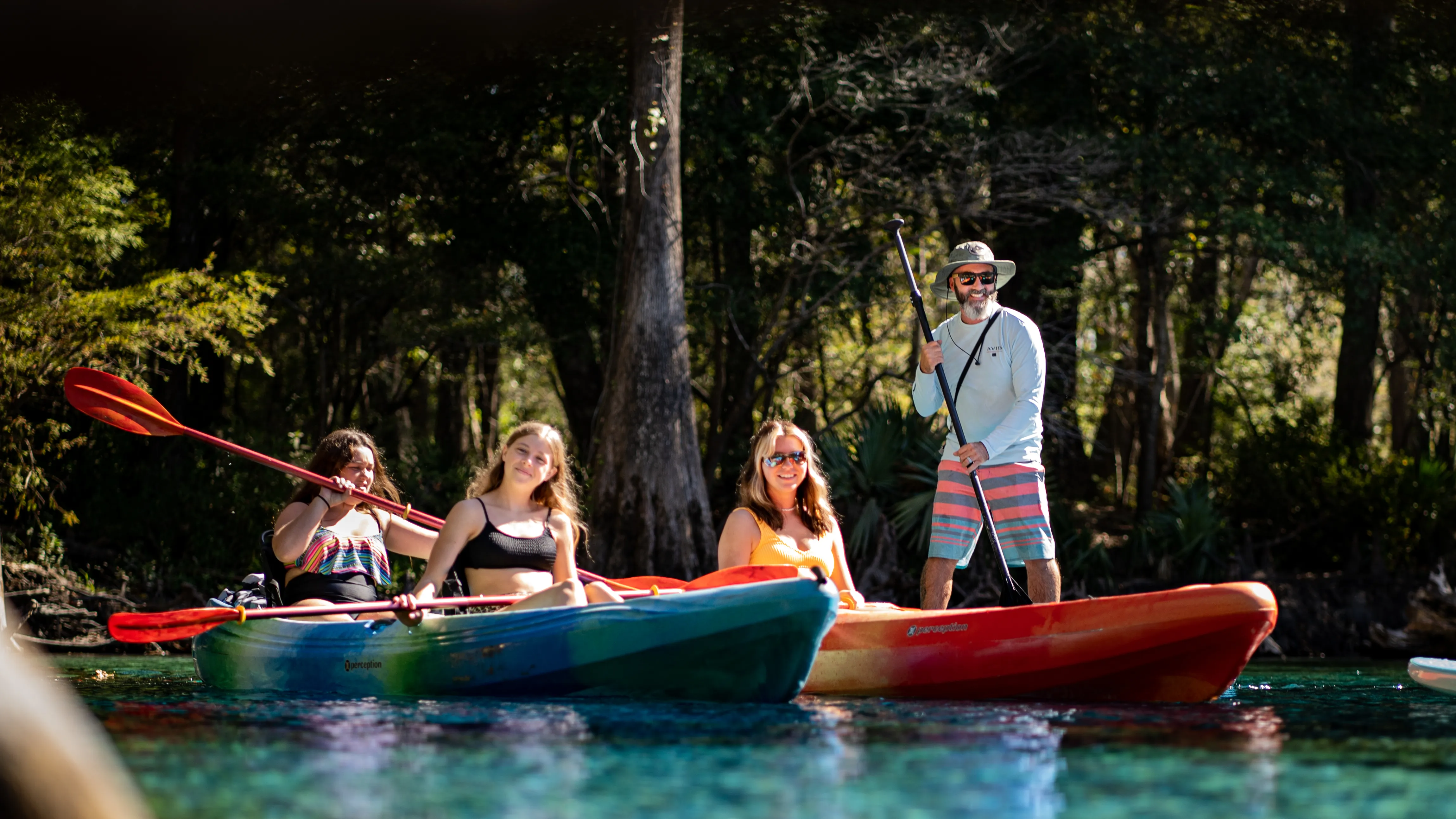 Two tandem kayaks and a paddleboarder pause together in turquoise spring water, the group smiling for the camera beneath towering cypress trees
