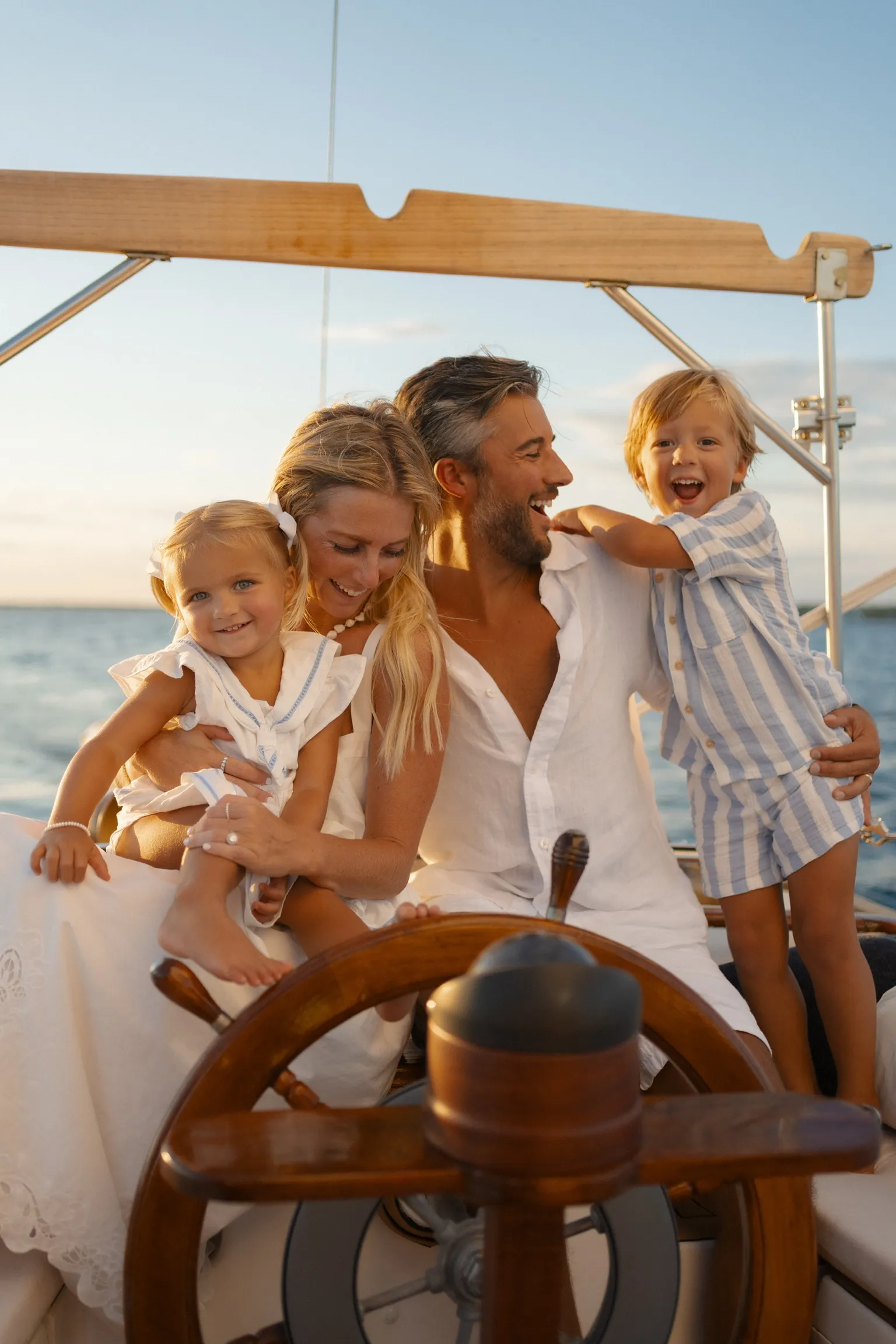 Two parents and two young children laughing together at the wheel of a sailboat at sunset